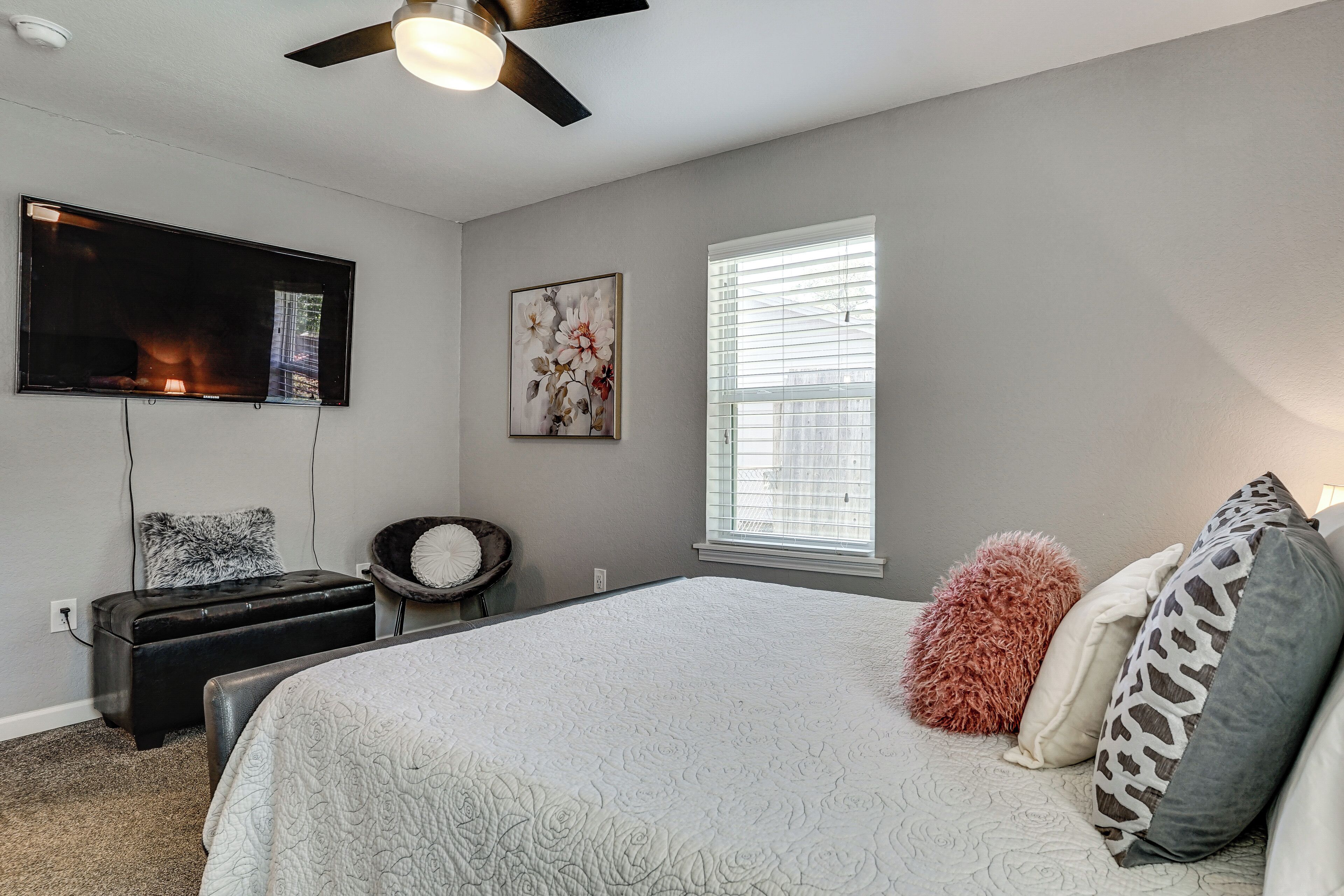 A bedroom with a bed, a TV mounted on the wall, a window with blinds, and a ceiling fan.