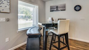 A dining area with a table, chairs, a bench, a window with blinds, and wall art including a compass and a lighthouse painting. - 2 Mi to Stadiums & UT Arlington! Stylish Home (Arlington)