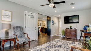 A living room with a ceiling fan, a TV, a patterned rug, and a door leading to another room. - 2 Mi to Stadiums & UT Arlington! Stylish Home (Arlington)