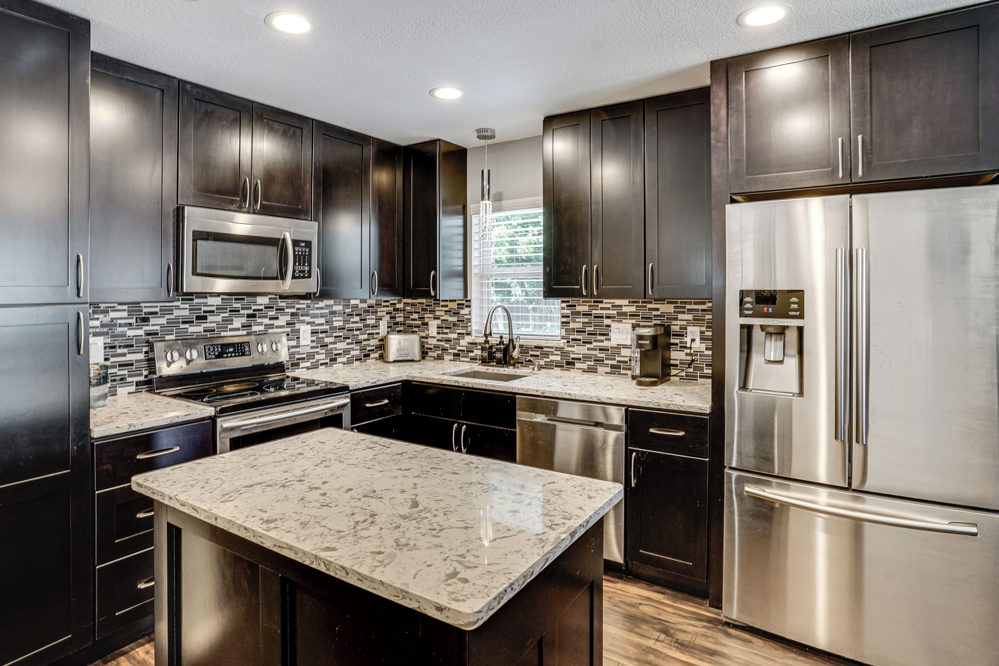 A modern kitchen with dark cabinetry, stainless steel appliances, a marble island, and a tiled backsplash.