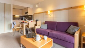 A modern hotel room with a purple sofa, a wooden coffee table with a teapot and cup, a dining area with chairs and a table, and a kitchenette. - Résidence Pierre & Vacances Mountain view (Flaine)