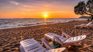 Ein Strand mit weißen Liegestühlen und einem Tisch, ein Sonnenuntergang im Hintergrund und ein Baum zur rechten Seite.