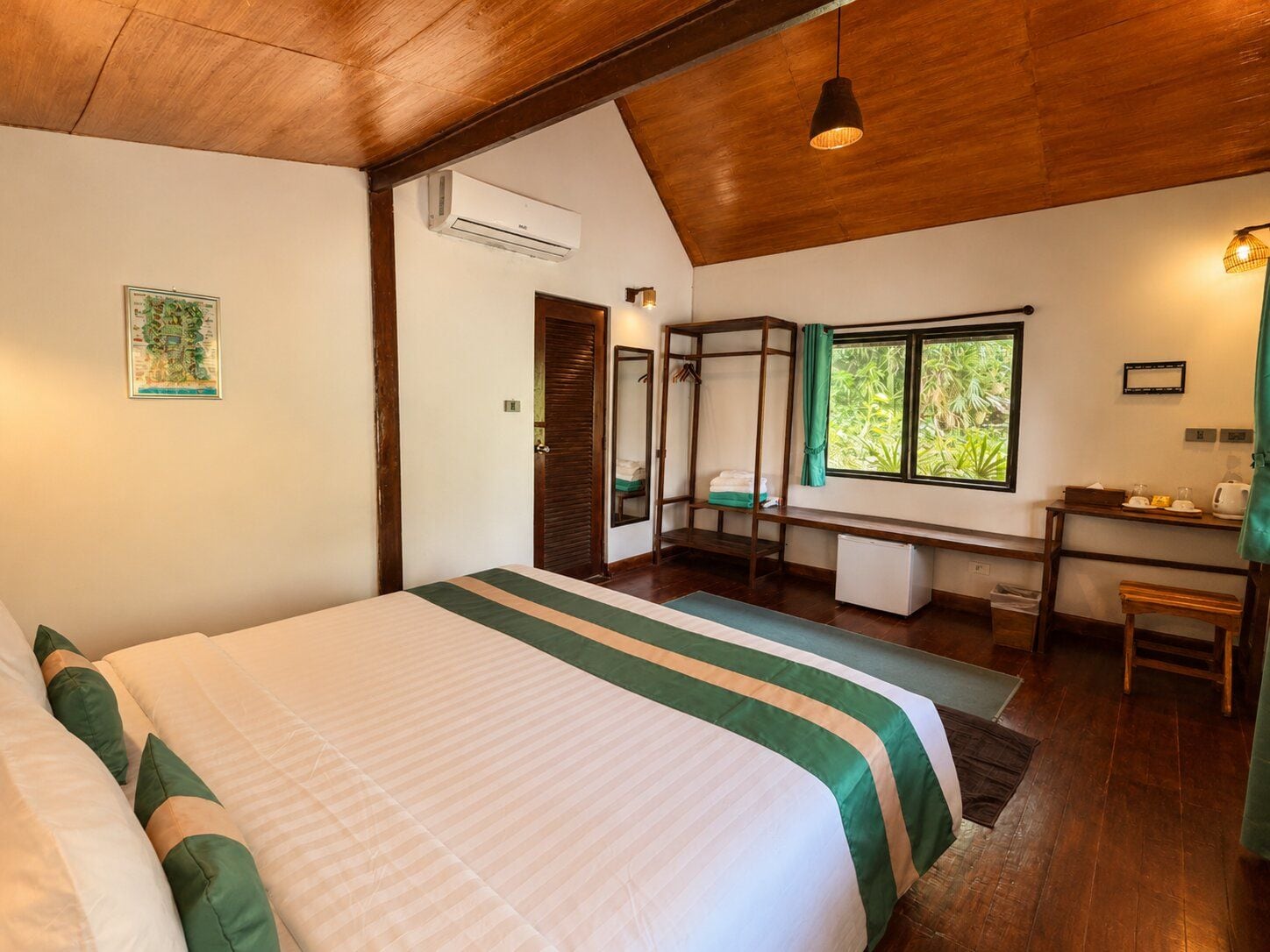A bedroom with a large bed, a desk, and a window with a view of greenery.