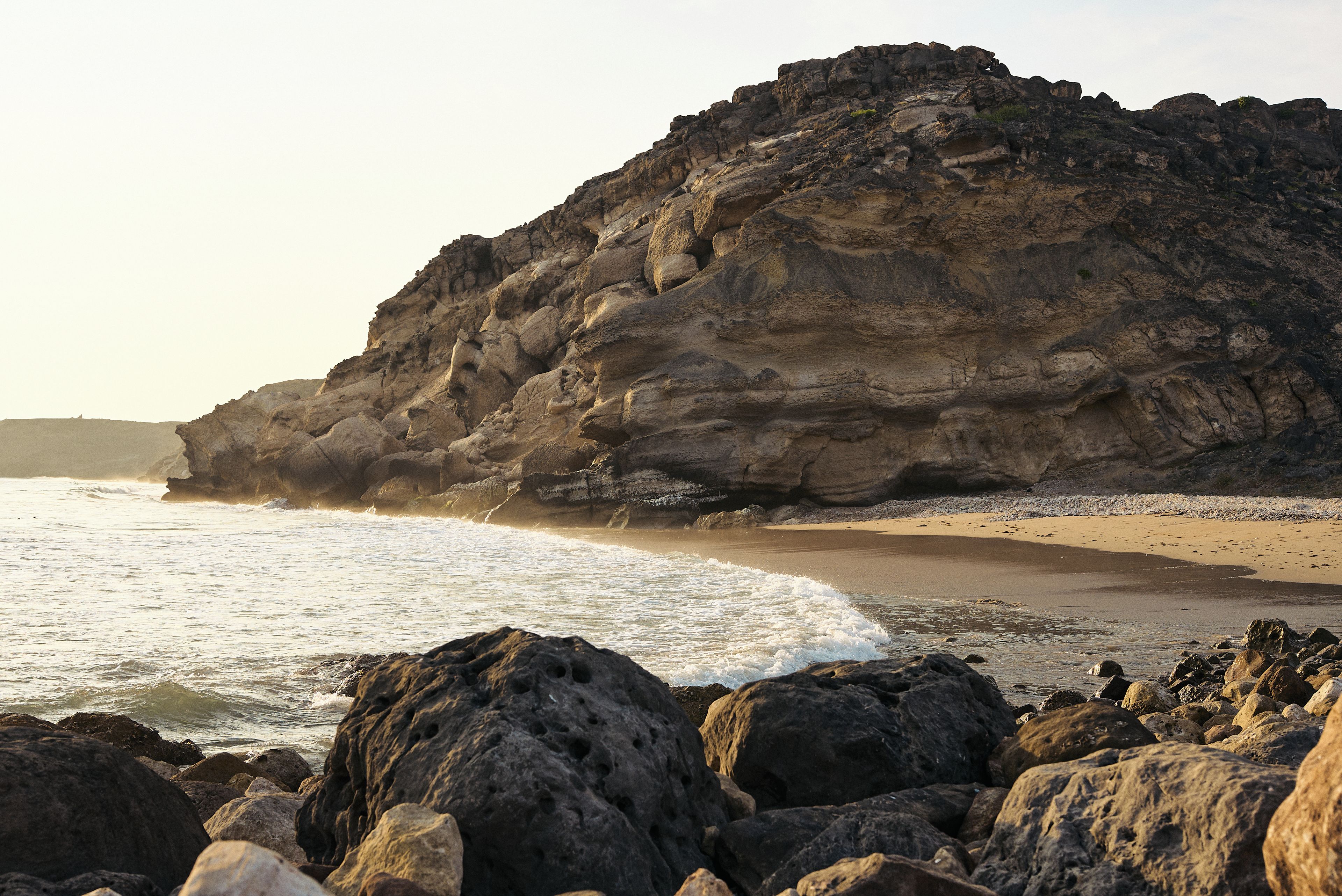 Costa rochosa com um penhasco imponente e ondas quebrando na praia.