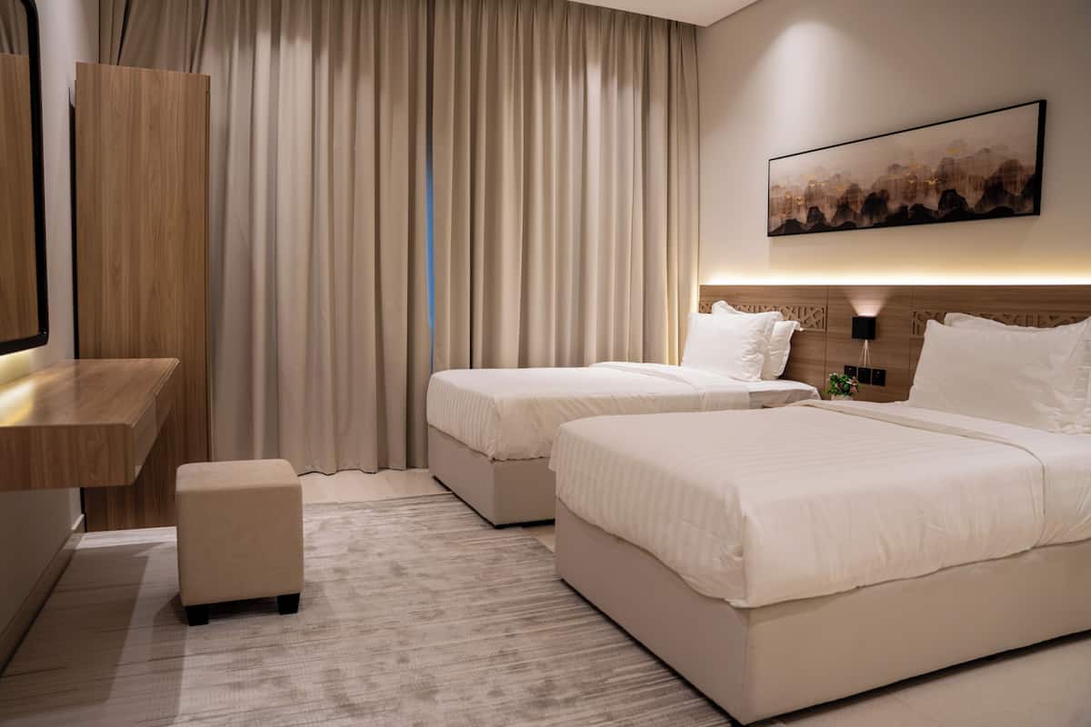 A hotel room with two beds, a desk, a chair, a small ottoman, and a painting on the wall.
