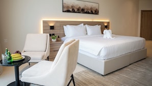 Una habitación de hotel moderna con una cama grande, una zona de estar con una silla y una mesita, y una obra de arte colgada en la pared.