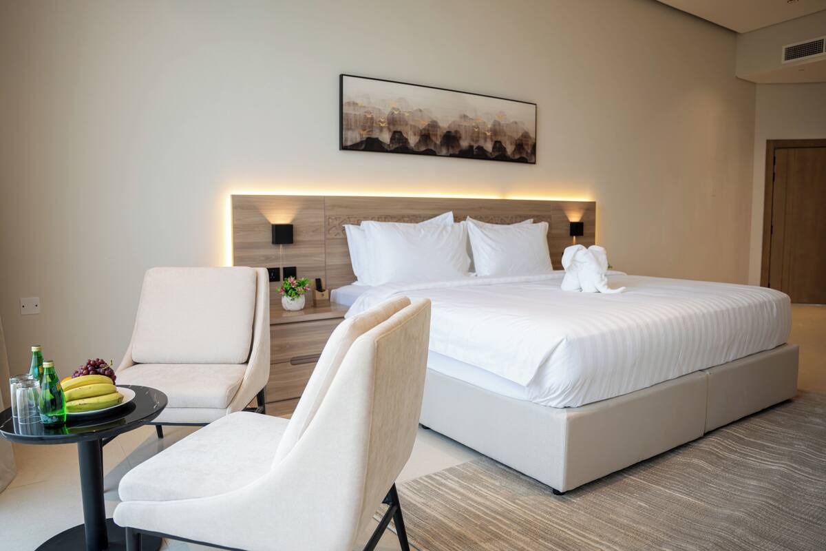 A modern hotel room with a large bed, a seating area with a chair and a small table, and a wall-mounted artwork.
