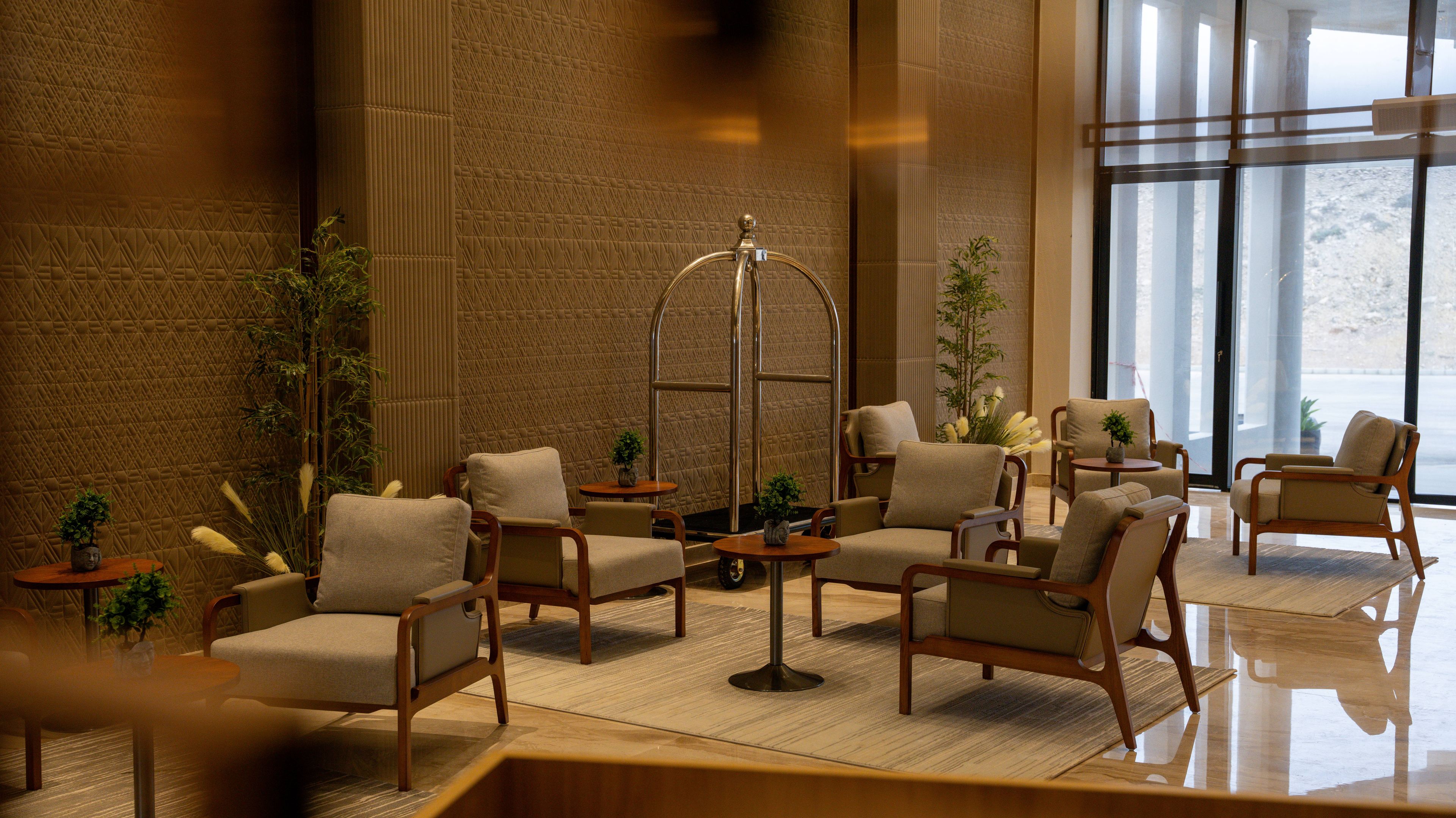 A hotel lobby with a seating area featuring armchairs and a high table, a large window providing a view, and a textured wall.
