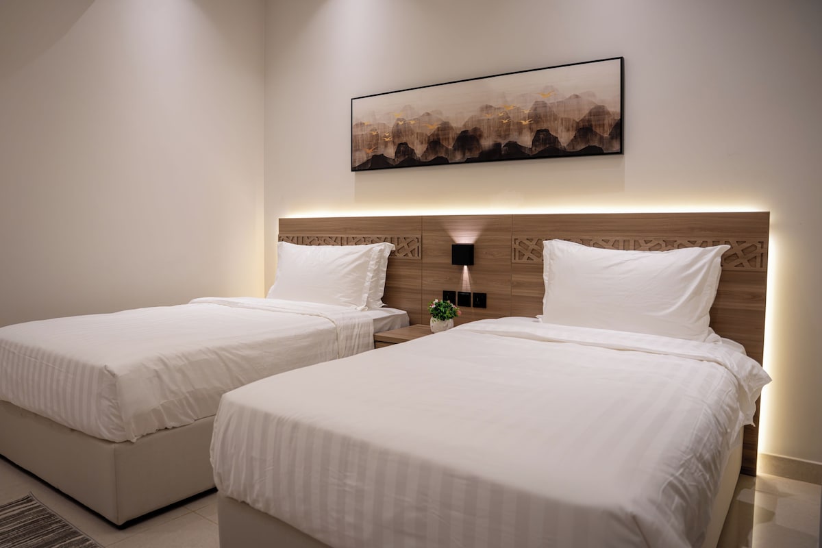 A hotel room with two beds, a nightstand, a wall-mounted lamp, and a painting.