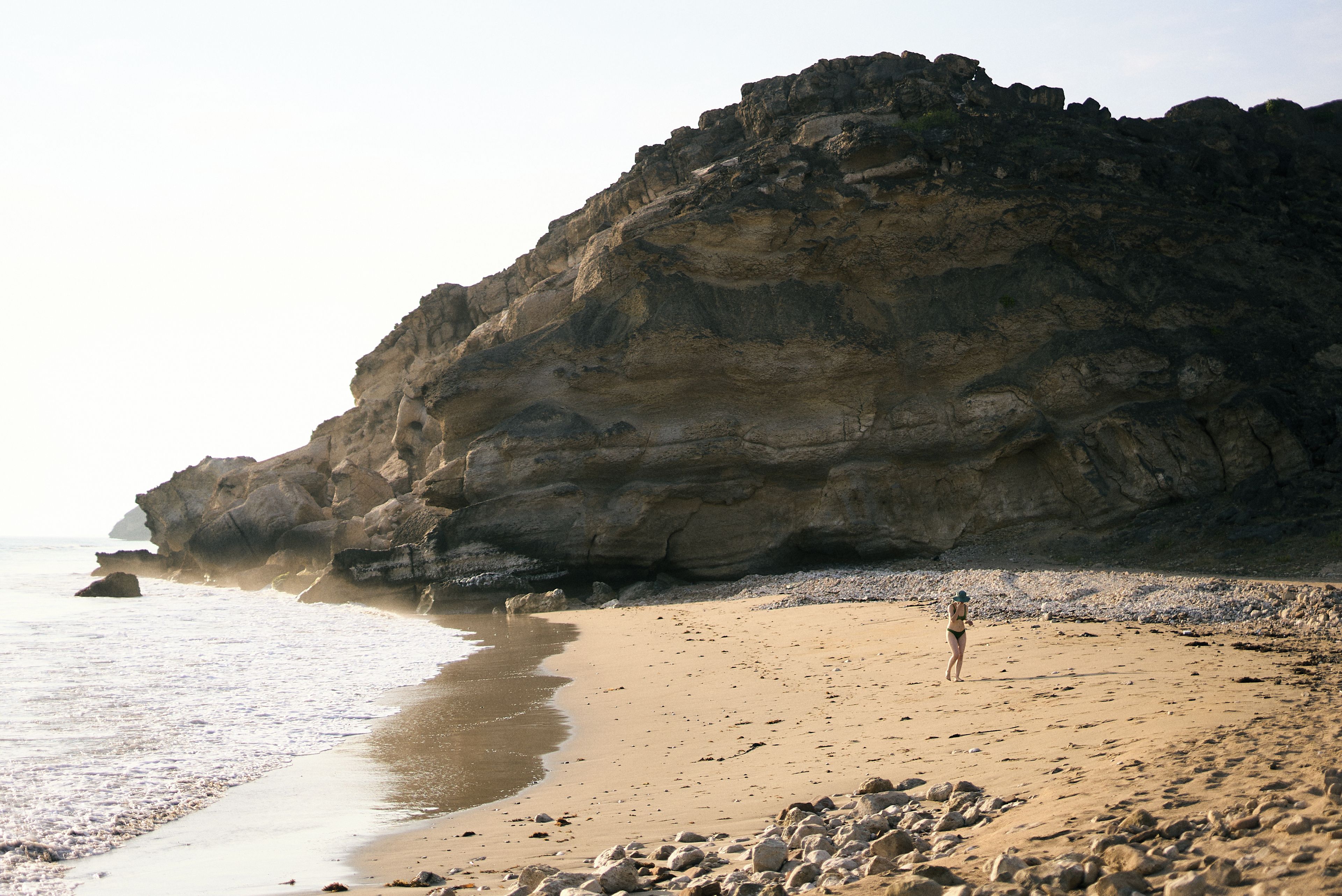 A beach with a rocky cliff and a person walking on the sand.