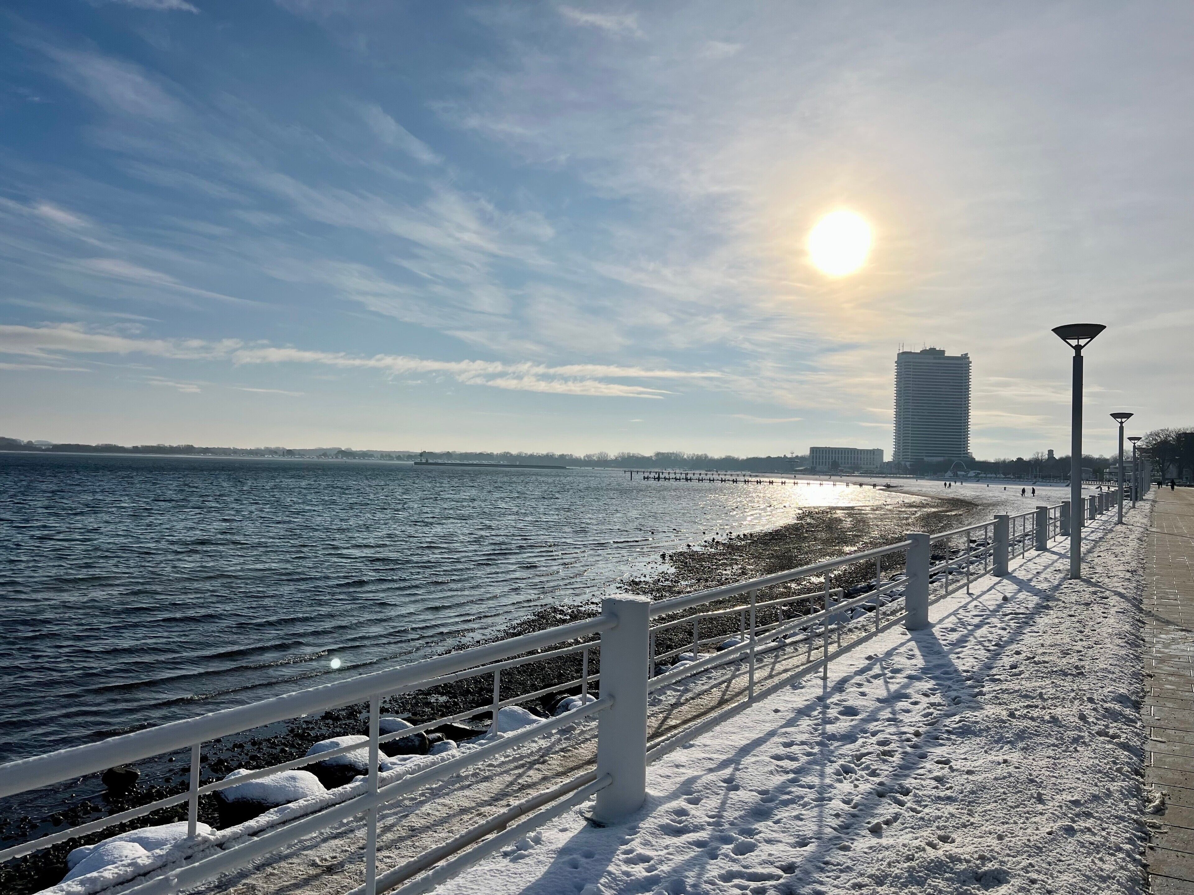 Une promenade en bord de mer avec une rambarde en métal, de la neige au sol et un ciel dégagé.