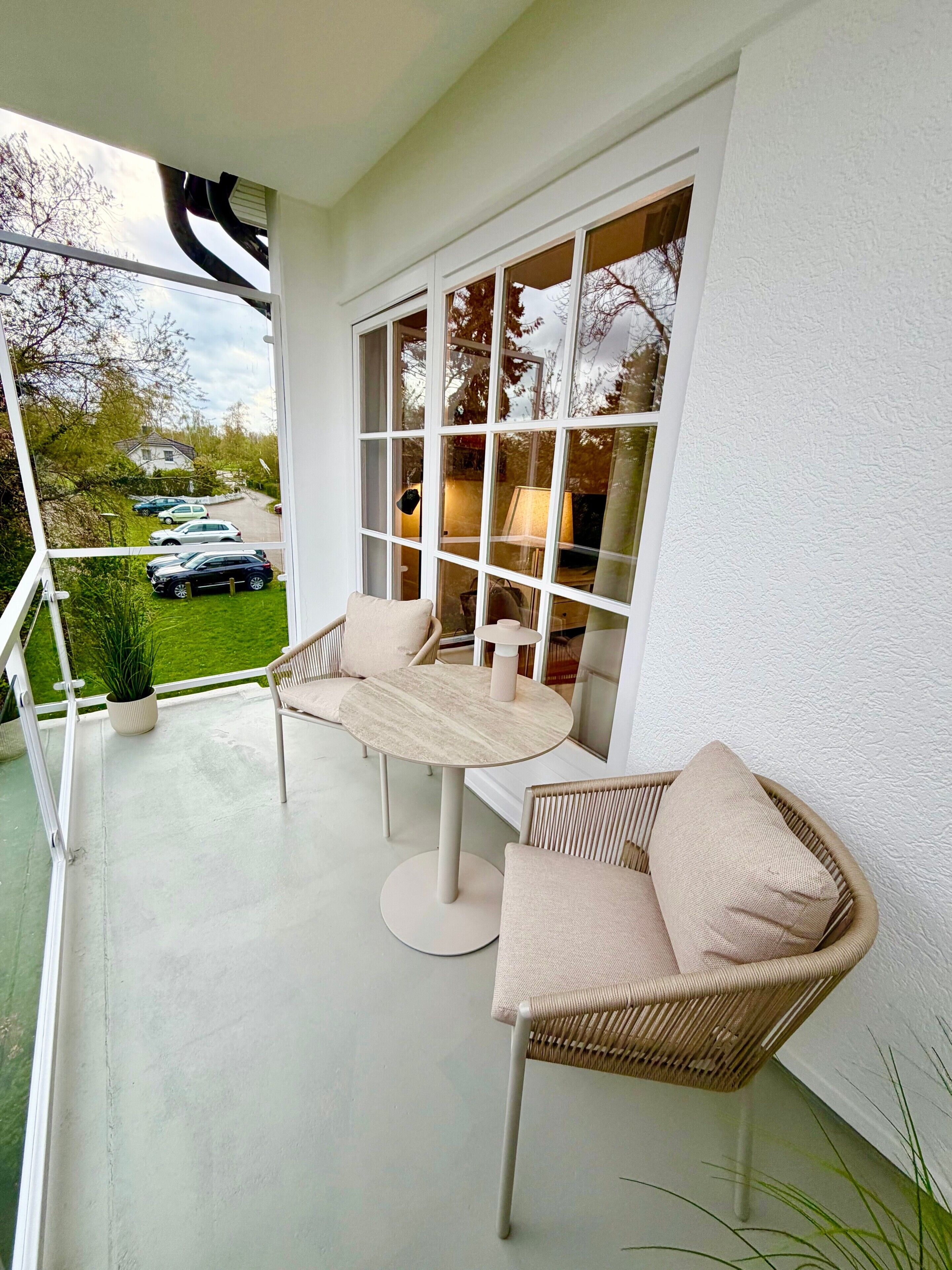 A balcony with a table and two wicker chairs, a potted plant, and a view of parked cars and trees.