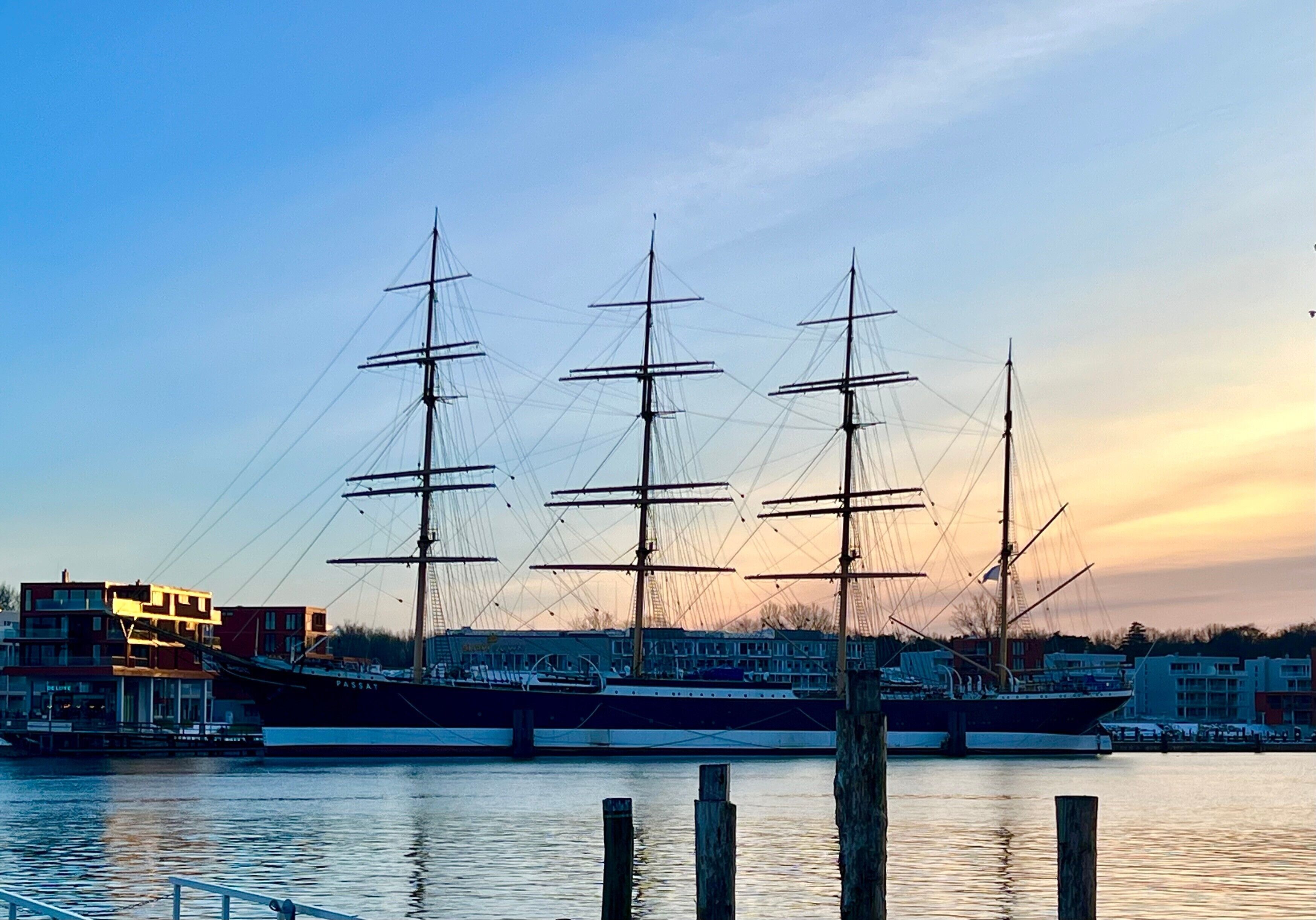 Ein großes Segelschiff liegt in einer Marina vor einer Gebäudereihe.