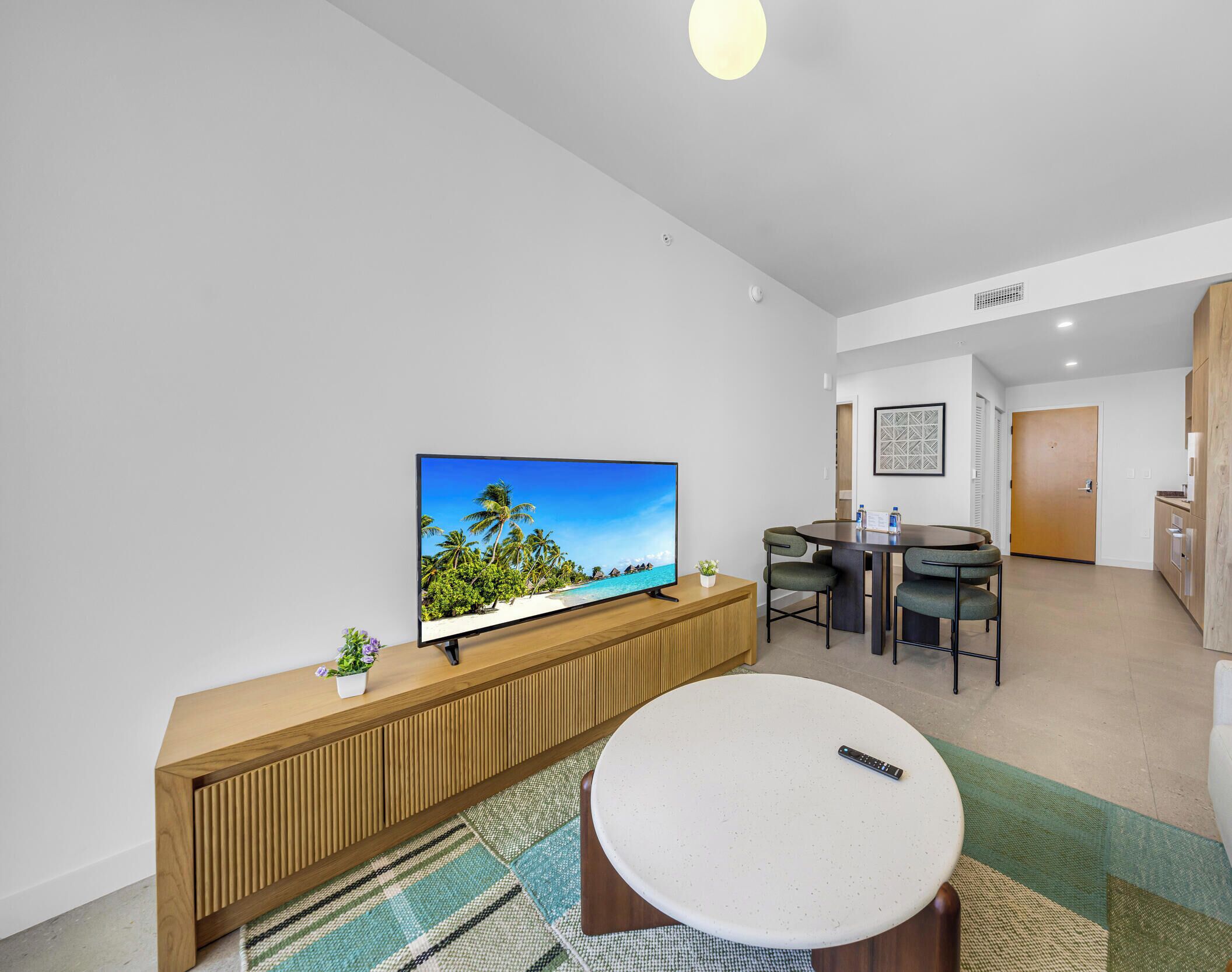 A modern living room with a flat-screen TV, a round coffee table, a dining area with a table and chairs, and a kitchen area with a wooden cabinet and a door.