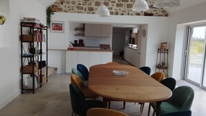 Een eetgedeelte met een houten tafel en kleurrijke stoelen, een keuken op de achtergrond, en een stenen muur.