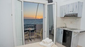 A compact kitchen with a sink, a stove, and a small refrigerator. There is a dining area with a table and chairs, and a view of the sea through a glass door.