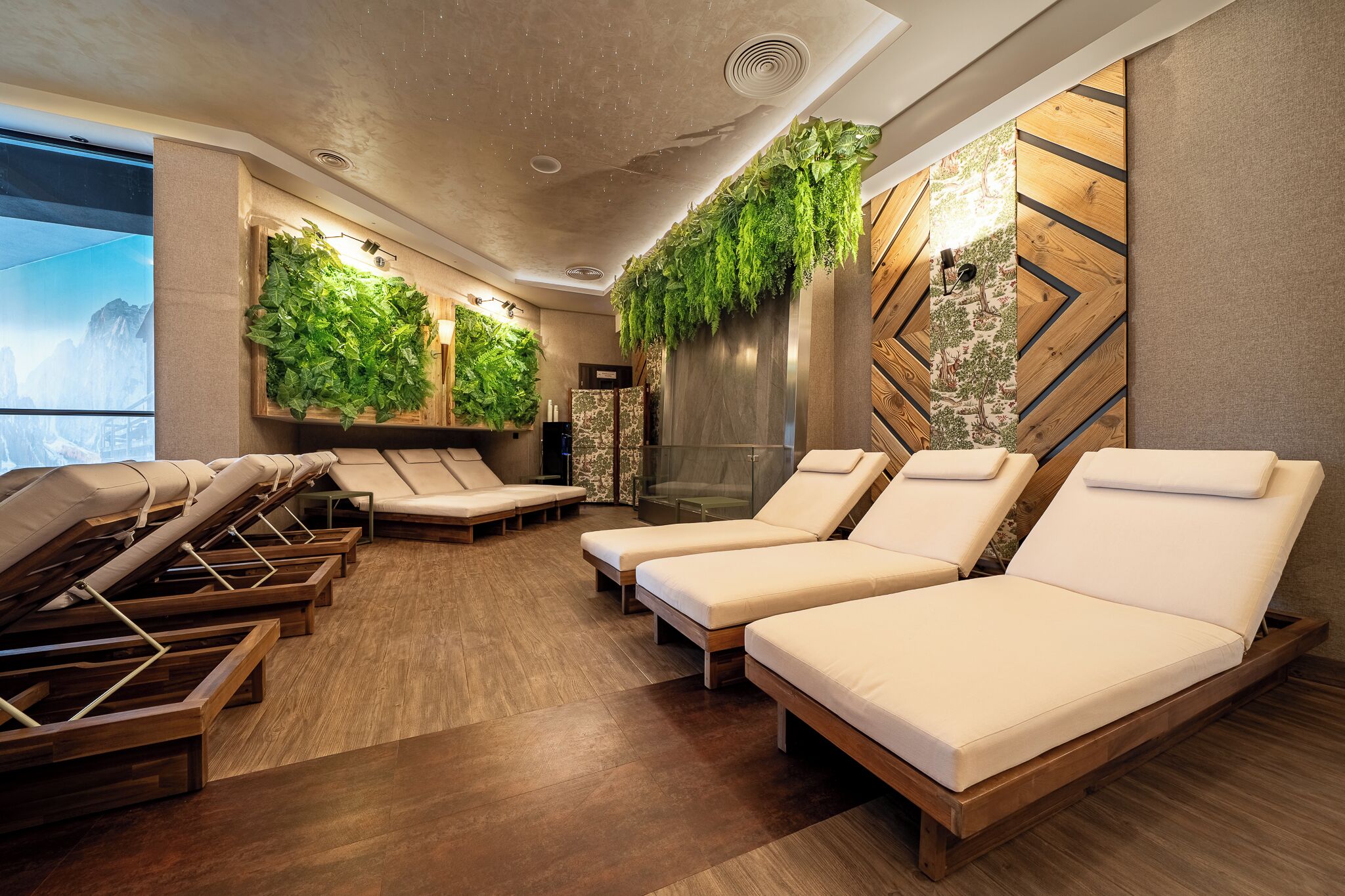A spa room with wooden flooring, multiple sun loungers, a wall with a vertical garden, and a large window with a mountain view.