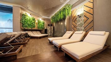 A spa room with wooden flooring, multiple sun loungers, a wall with a vertical garden, and a large window with a mountain view.