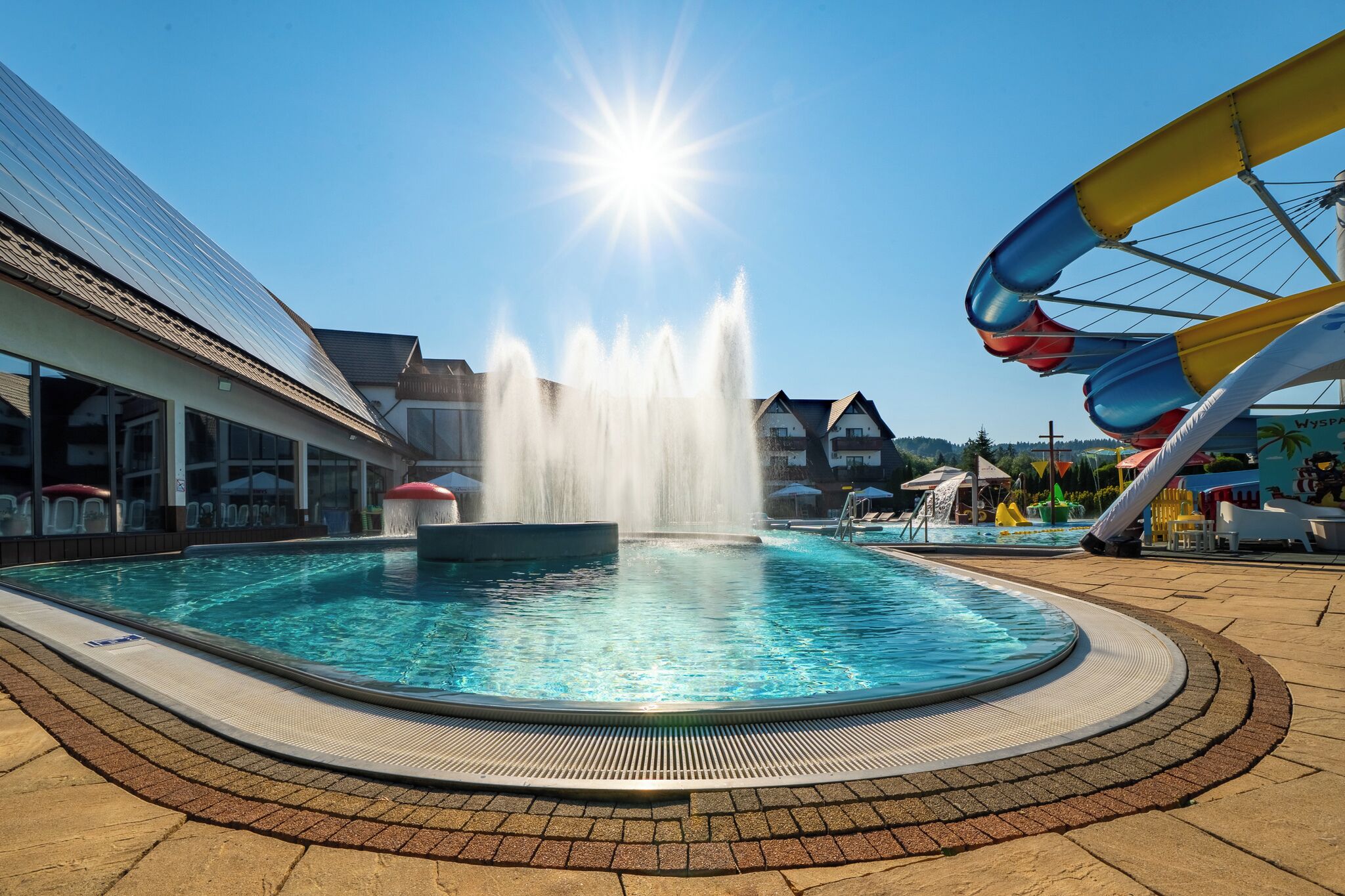 Una piscina con una fuente, un tobogán acuático y un edificio con techo inclinado.