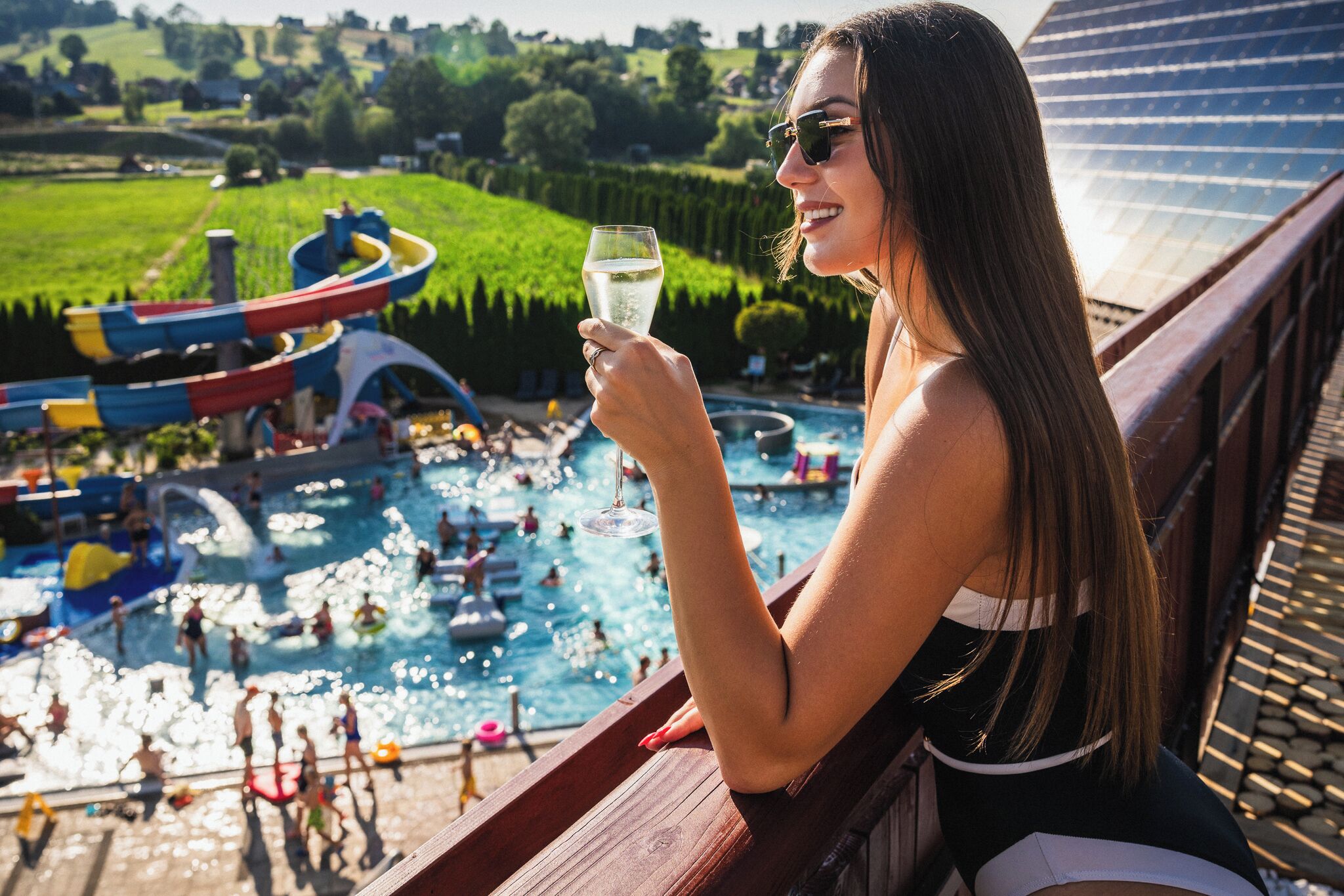 A person holding a glass of water, overlooking a swimming pool with people and water slides.