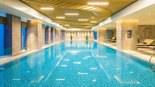 A large indoor swimming pool with a tiled floor, surrounded by marble walls and columns, and a ceiling with a wavy design.