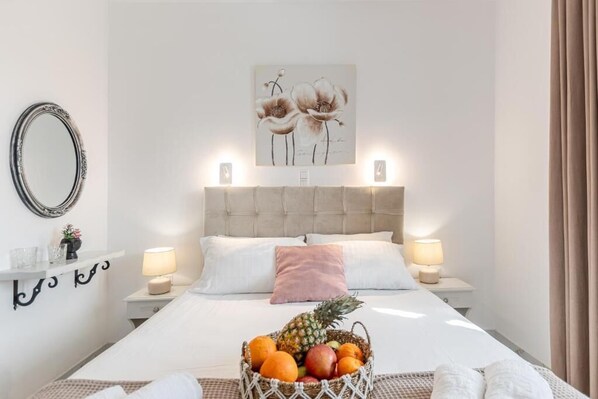 A hotel room with a bed, bedside lamps, a fruit basket, a wall-mounted mirror, and a floral artwork. - BY THE SEA (Agia Anna)