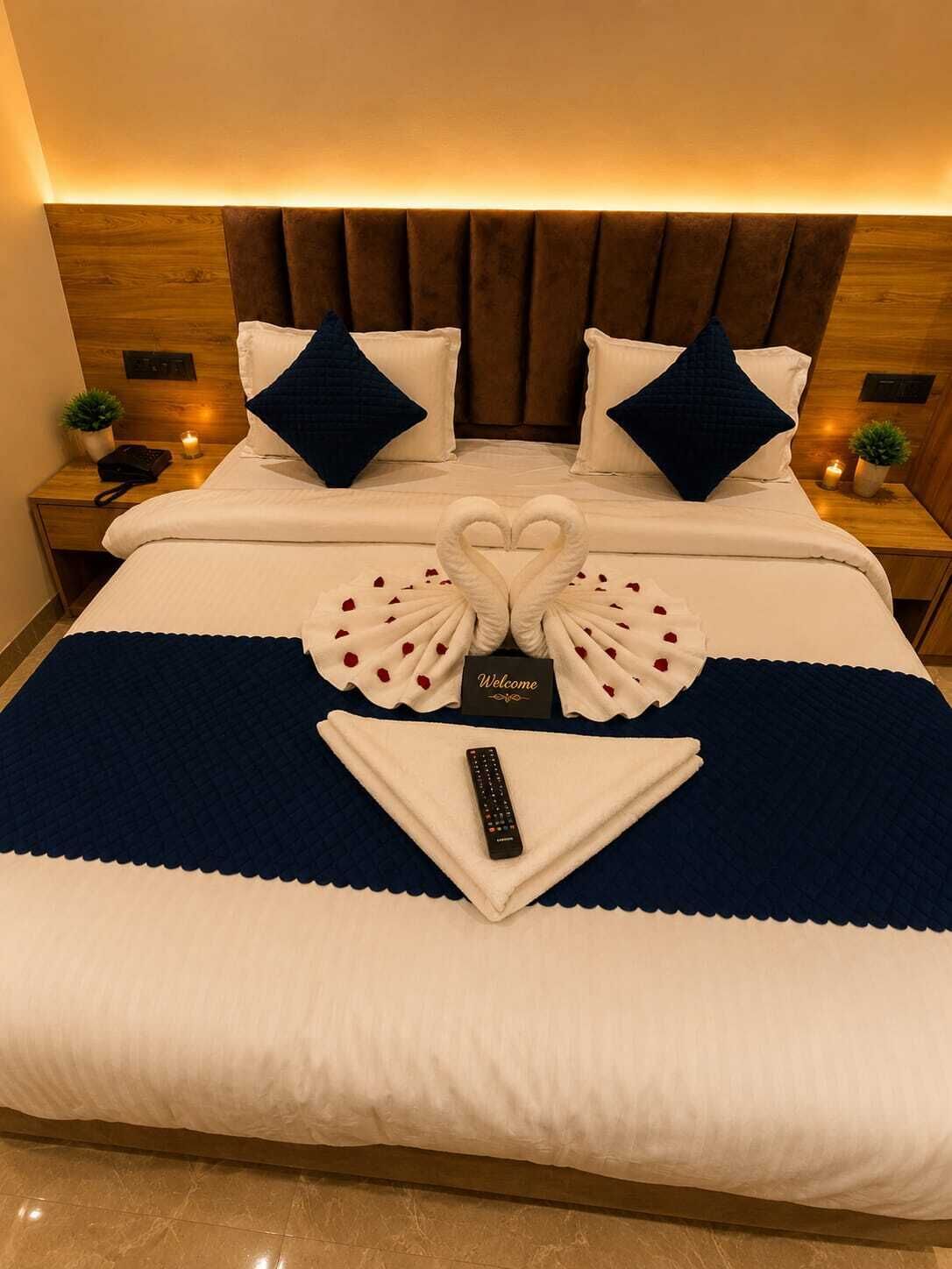 A neatly made bed with a heart-shaped towel arrangement, a welcome card, and a remote control on a blue and white bedspread.