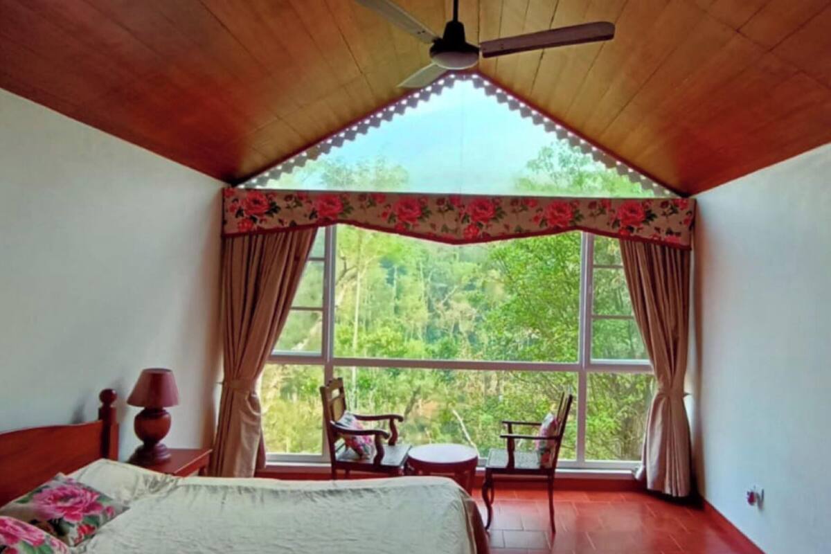 Un dormitorio con una ventana grande, ventilador de techo, cama, una silla y una mesita.