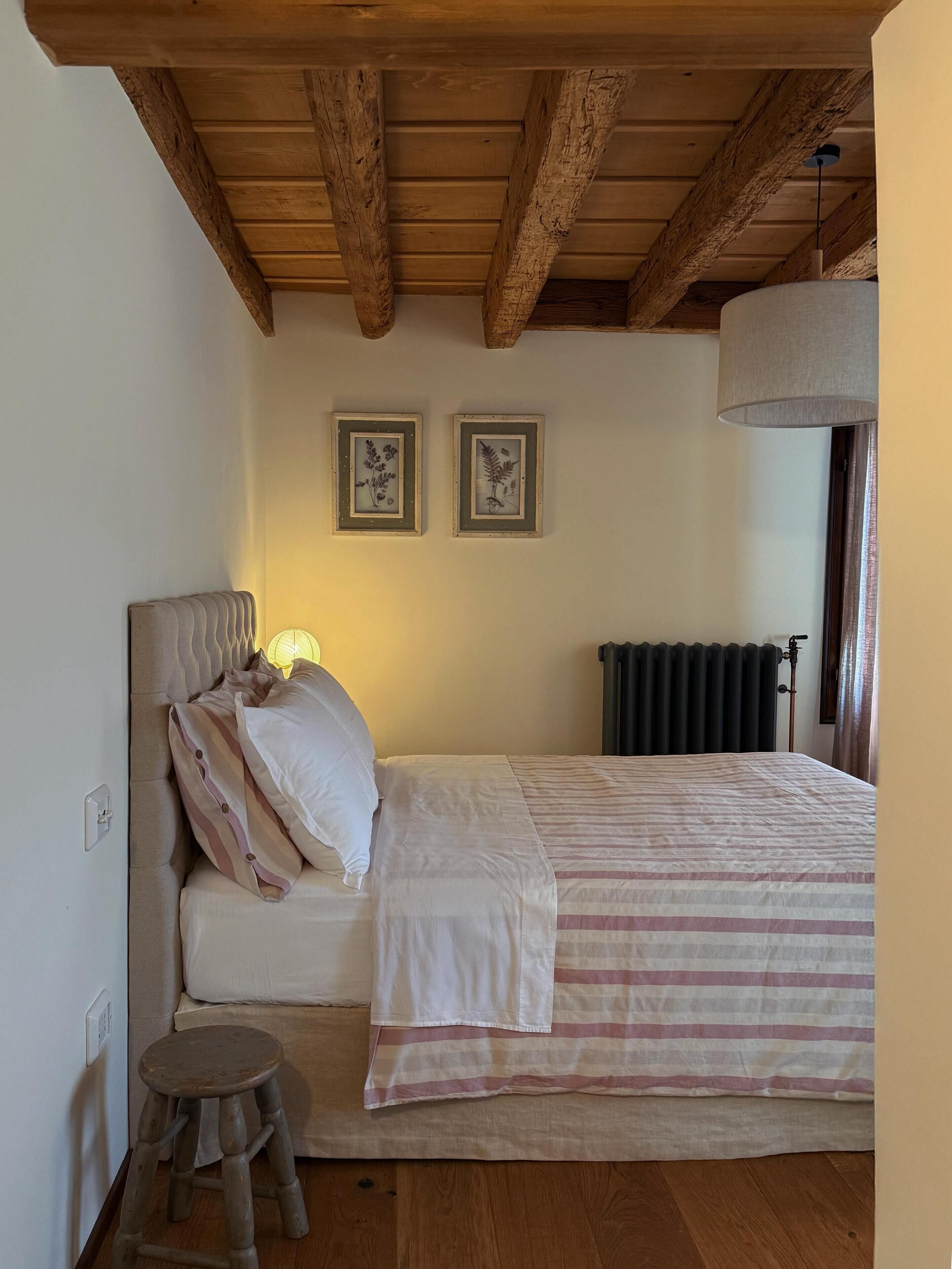 A bedroom with a wooden ceiling, a bed with a striped bedspread, a nightstand, a stool, and two framed botanical prints on the wall.
