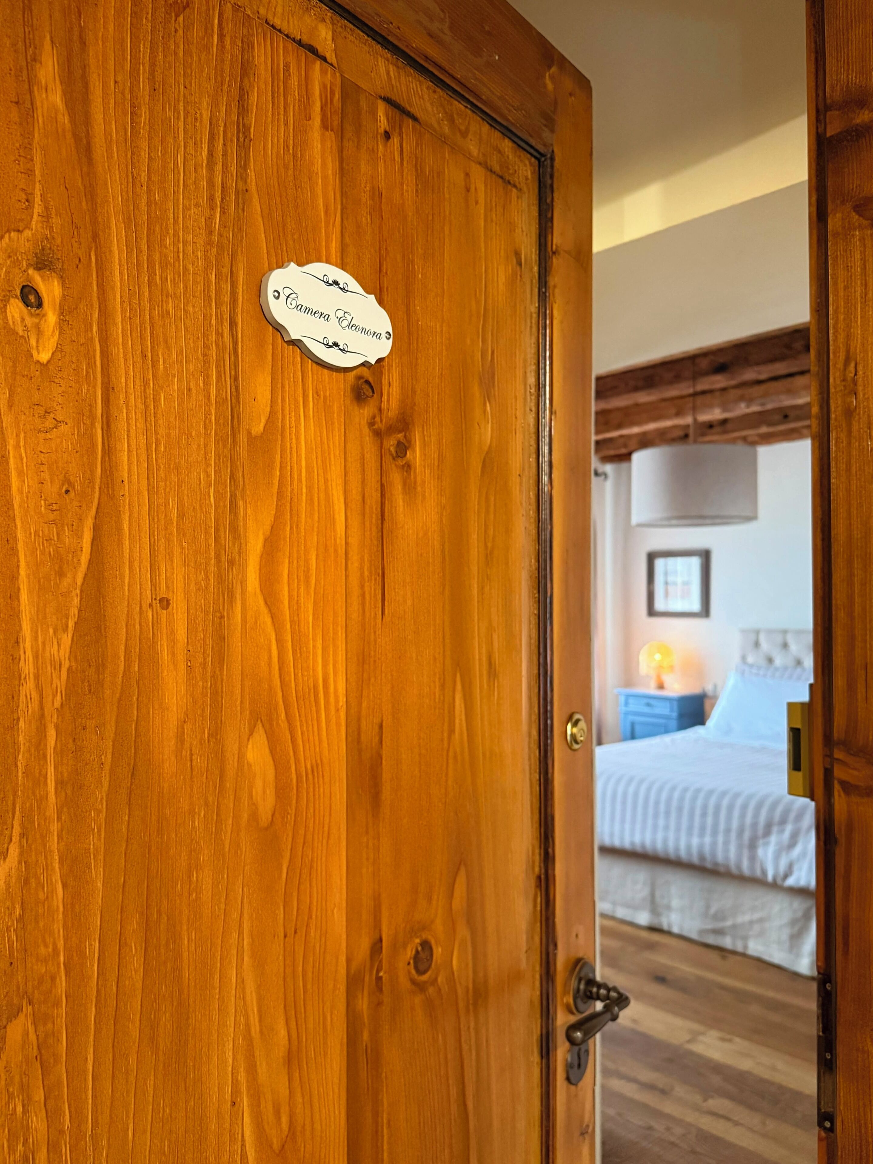Une porte en bois avec une plaque nominative, une chambre à coucher avec un lit, une table de chevet et une lampe.