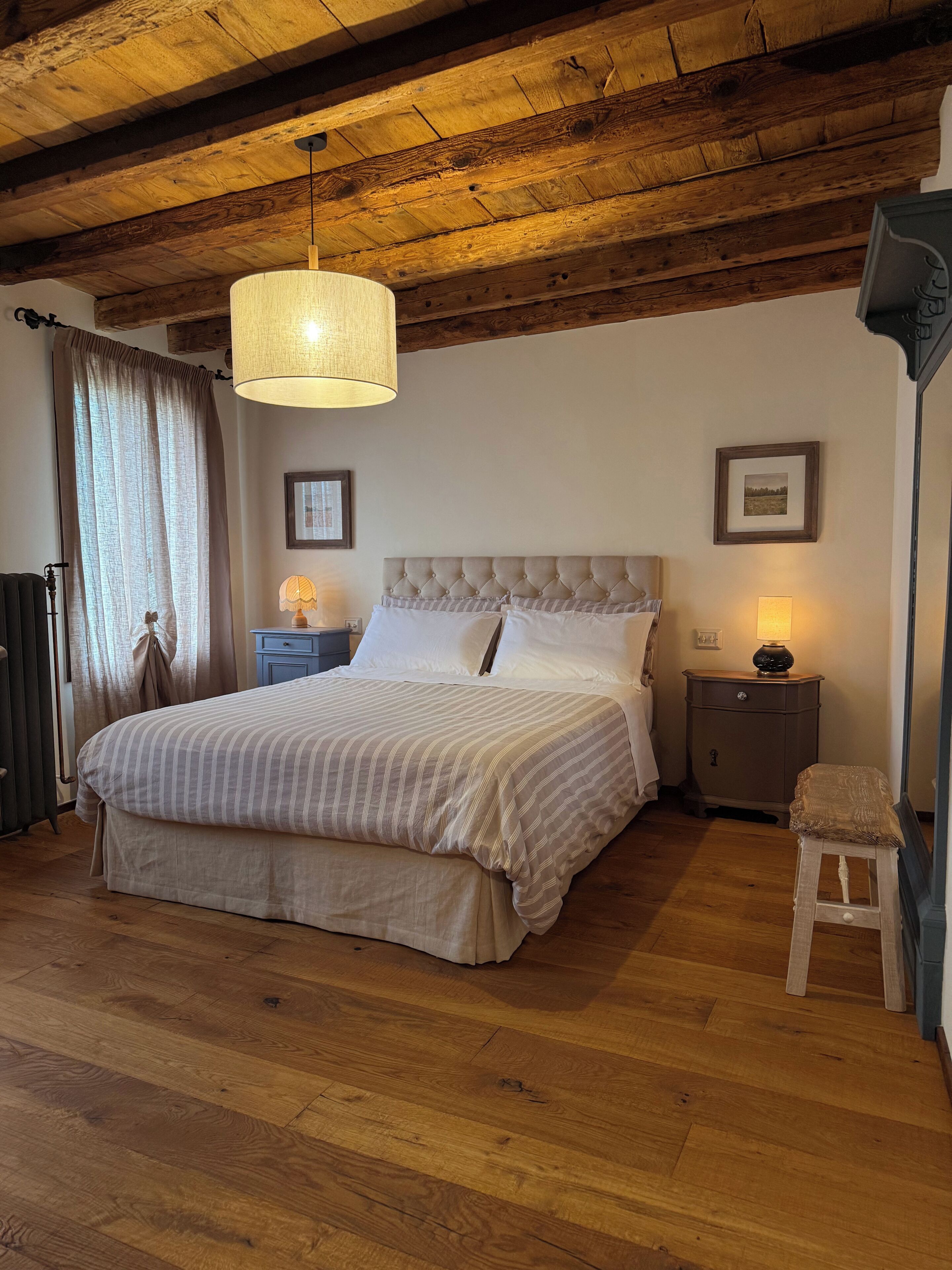 Un dormitorio con una cama grande, vigas de madera en el techo, una lámpara colgante, una mesita de noche con una lámpara y un cuadro en la pared.