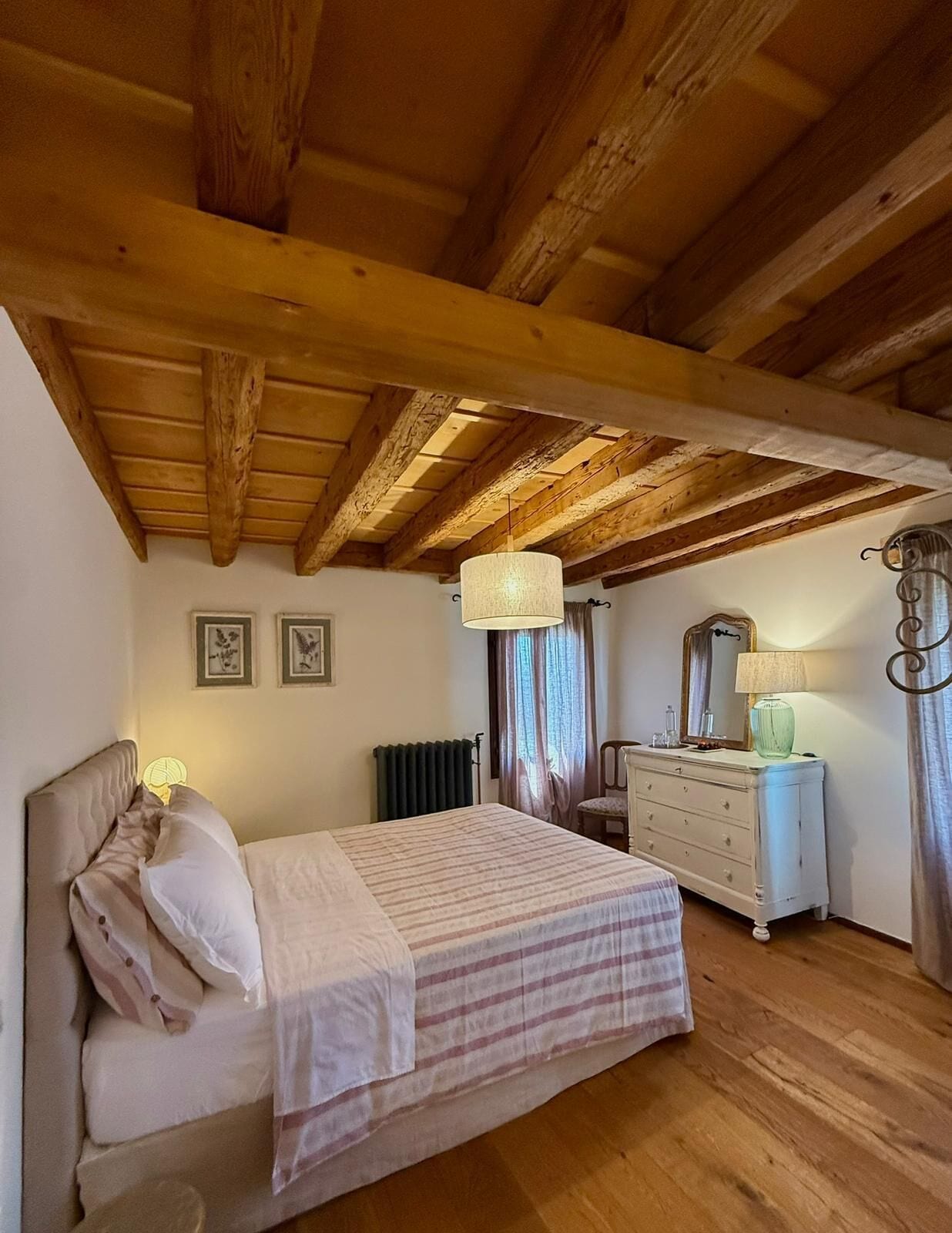 A bedroom with a wooden ceiling, a bed with a striped bedspread, a dresser, a mirror, and two wall-mounted picture frames.