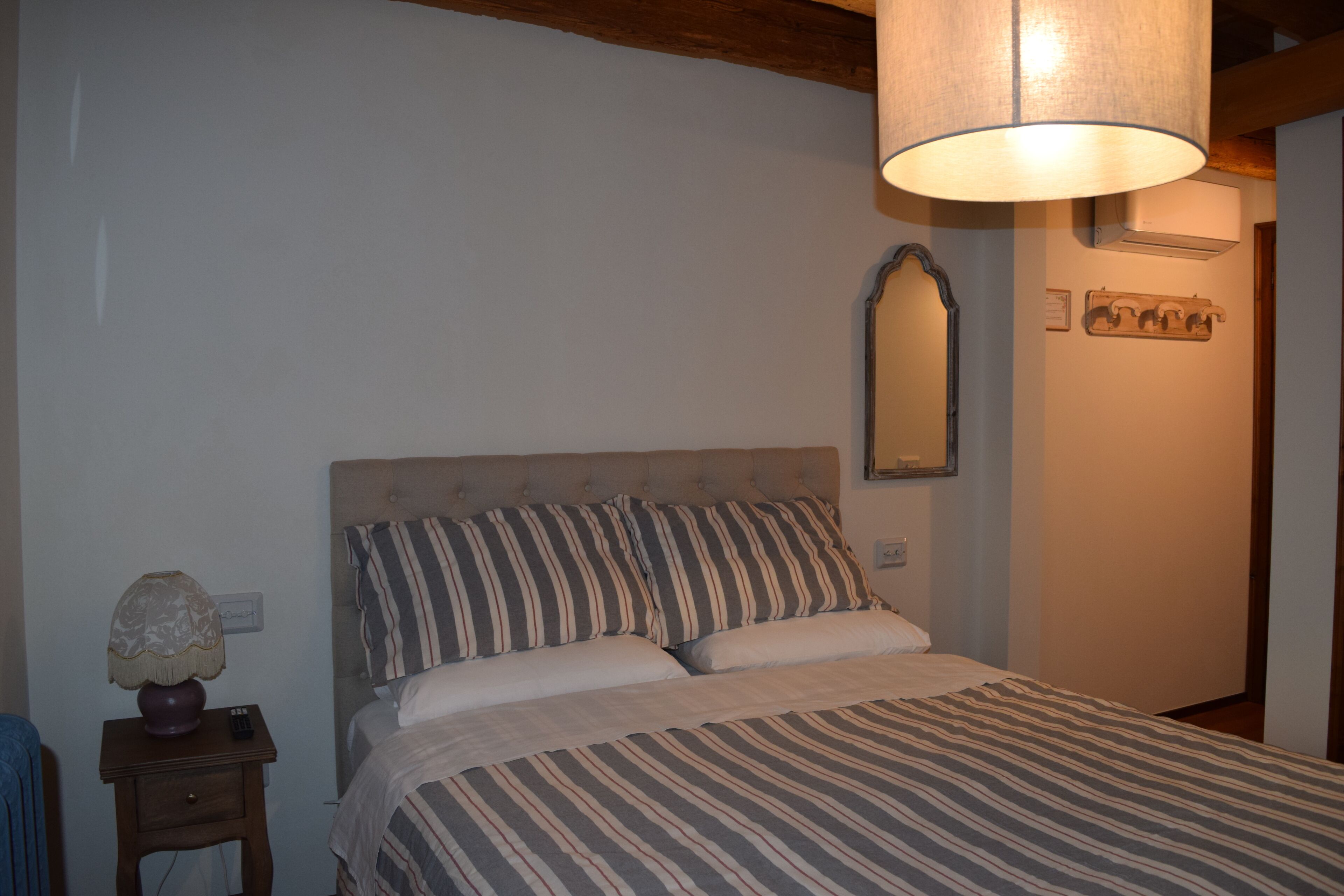 A bedroom with a bed, a nightstand, a lamp, a mirror, and a wall-mounted air conditioner.