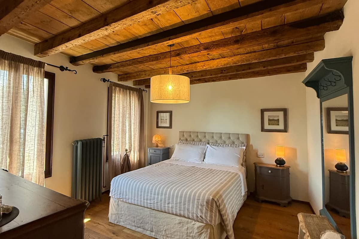 Un dormitorio con techo de madera, una cama con colcha a rayas, dos mesitas de noche con lámparas, un aparador y un espejo.