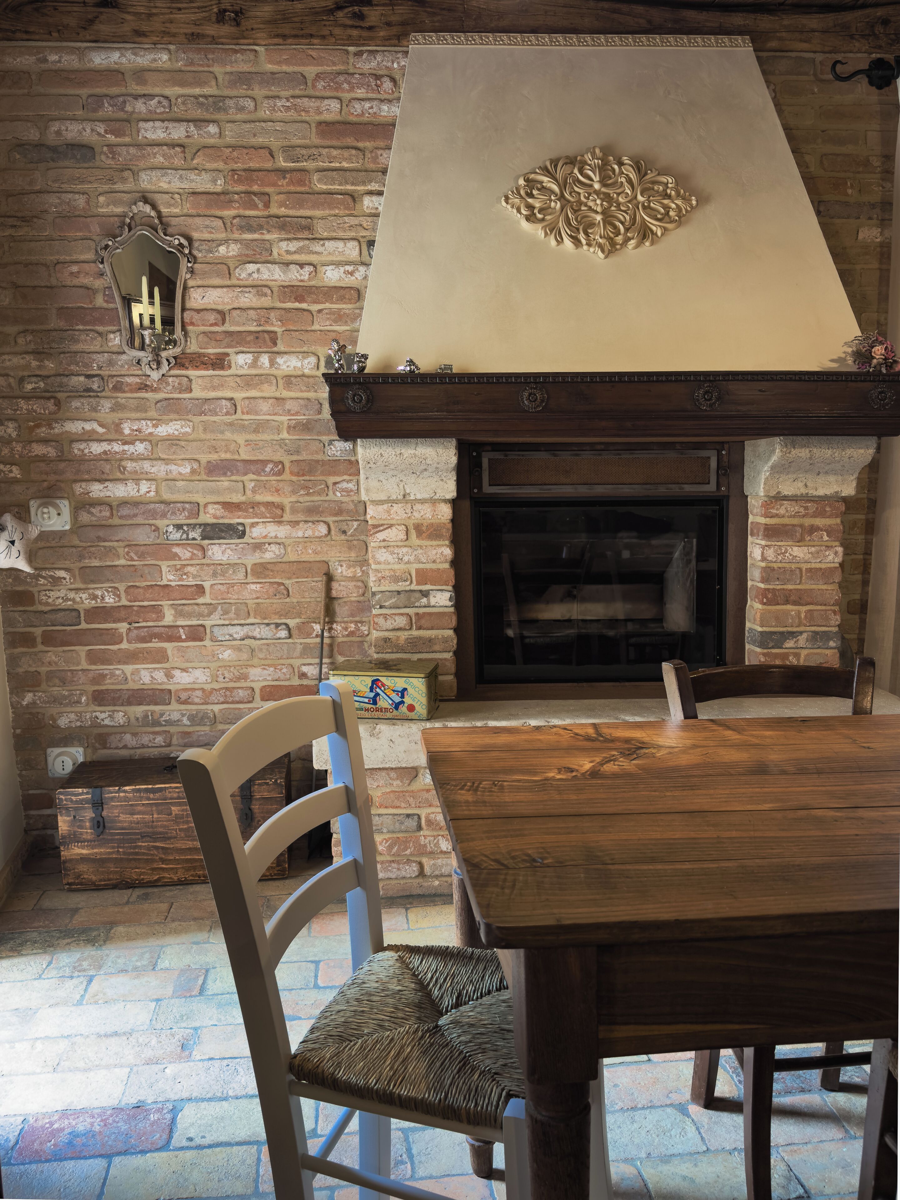Una chimenea de ladrillo con repisa, una mesa de madera y una silla de mimbre en una habitación con pared de ladrillo.