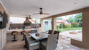 Patio esterno con tavolo e sedie, ventilatore a soffitto e vista su un giardino.