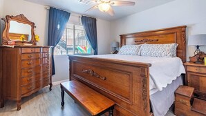 Un dormitorio con una cama de madera, un aparador, un banco, un ventilador de techo y una ventana con cortinas.