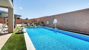 Una piscina rectangular con aro de baloncesto, rodeada por un muro de ladrillo y una zona cubierta de césped.