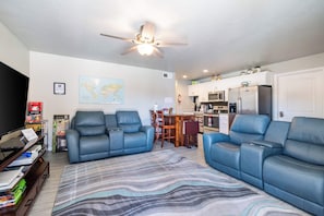 A living room with a sofa, a television, a ceiling fan, a map on the wall, and a kitchen in the background. - Grandma's Southern Comfort Vacation Rental (Hurricane)