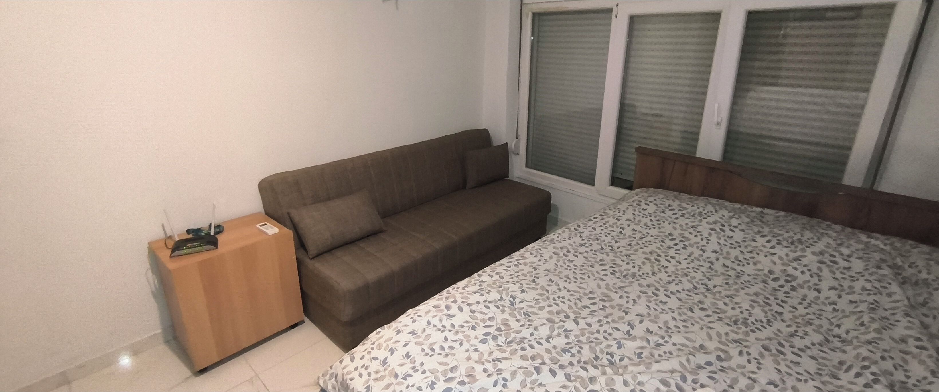 A room with a bed, a sofa, a wooden side table, and a window with closed blinds.