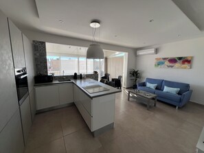 A modern kitchen with a central island, a dining area with a blue sofa, and a large window providing a view of the cityscape. - Luxury Penthouse 2-bedroom Jacuzzi  in Ix-Xagħra (Ix-Xagħra)