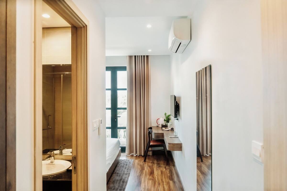 Una habitación de hotel con piso de madera, una cama, un escritorio con silla, un ventanal con cortinas y un baño con lavamanos y espejo.