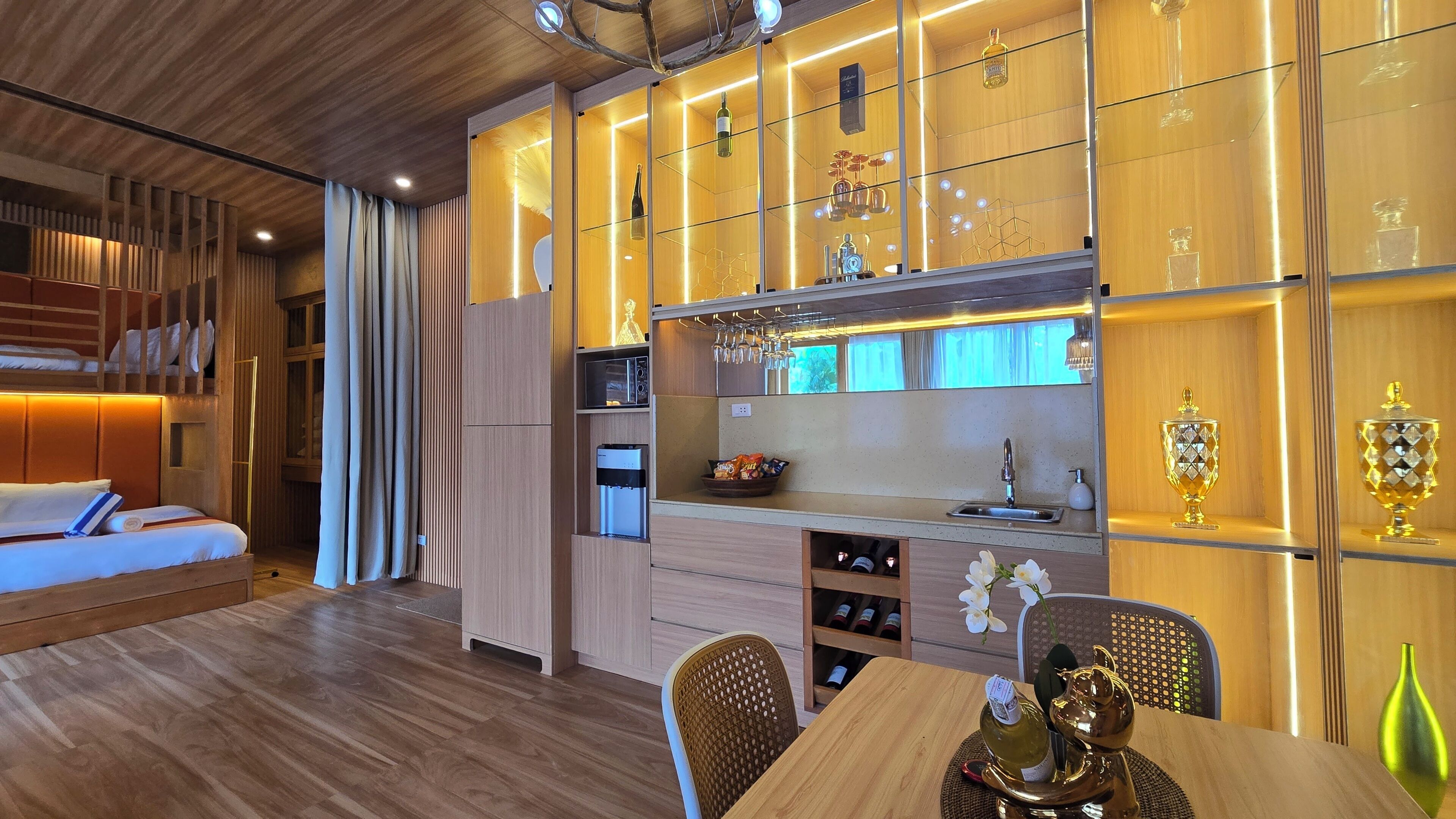 Quarto de hotel moderno com beliche, área de refeições com mesa e cadeiras, cozinha com pia e espaço para armazenamento de vinho, e um vaso decorativo na mesa.