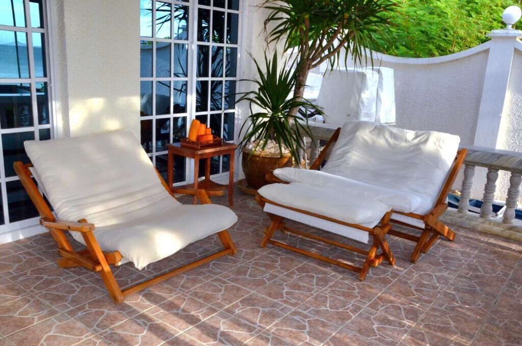 Deux fauteuils de jardin en bois, dotés de coussins blancs, sont placés sur une terrasse carrelée.