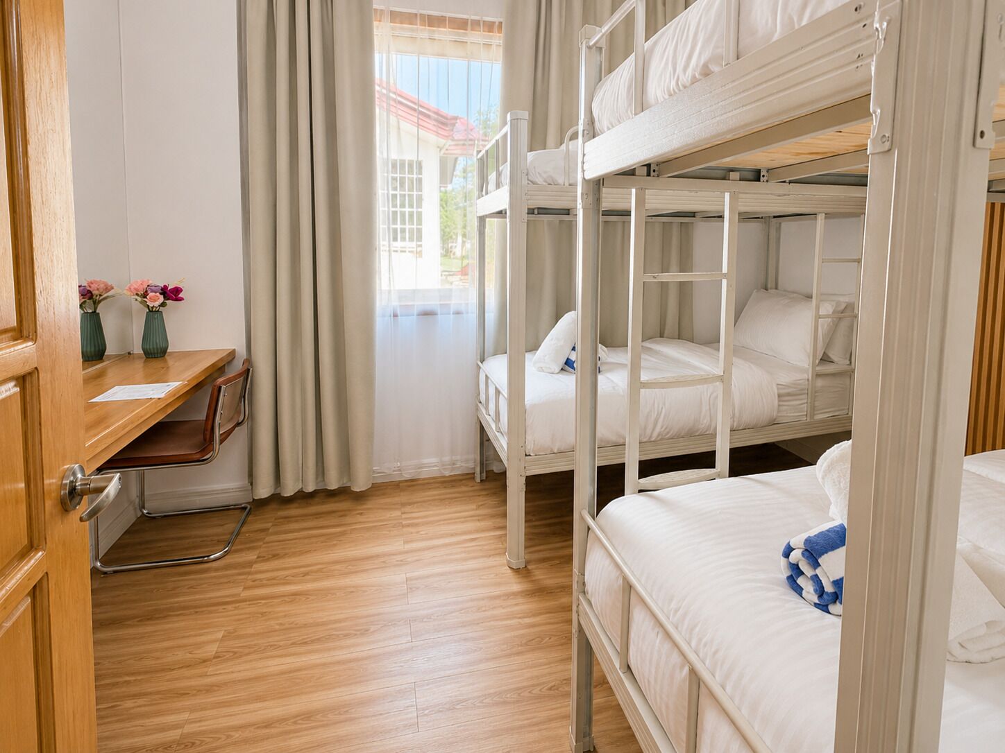 Ein Schlafraum mit Etagenbetten, einem Holztisch und Stühlen, einem Fenster mit Vorhängen und Blick auf ein Gebäude.