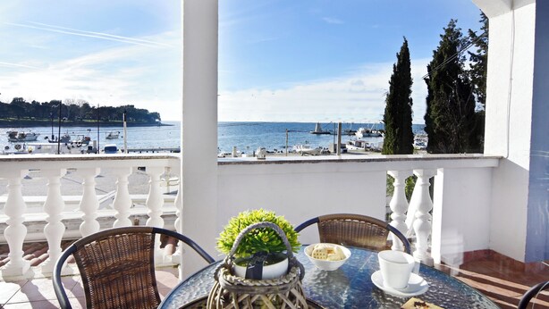 Un balcon avec vue sur la mer, des fauteuils en osier, une table avec un plateau en verre et une plante en pot.