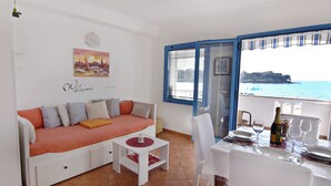 Une chambre meublée avec un canapé, une table à manger dressée pour deux personnes, une fenêtre donnant sur la mer et une pancarte « Bienvenue » accrochée au mur.
