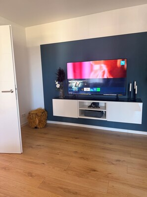 Ein modernes Wohnzimmer mit einem flachbildfernseher an einer weißen Wand, Holzboden und einem kleinen Zierhocker.
