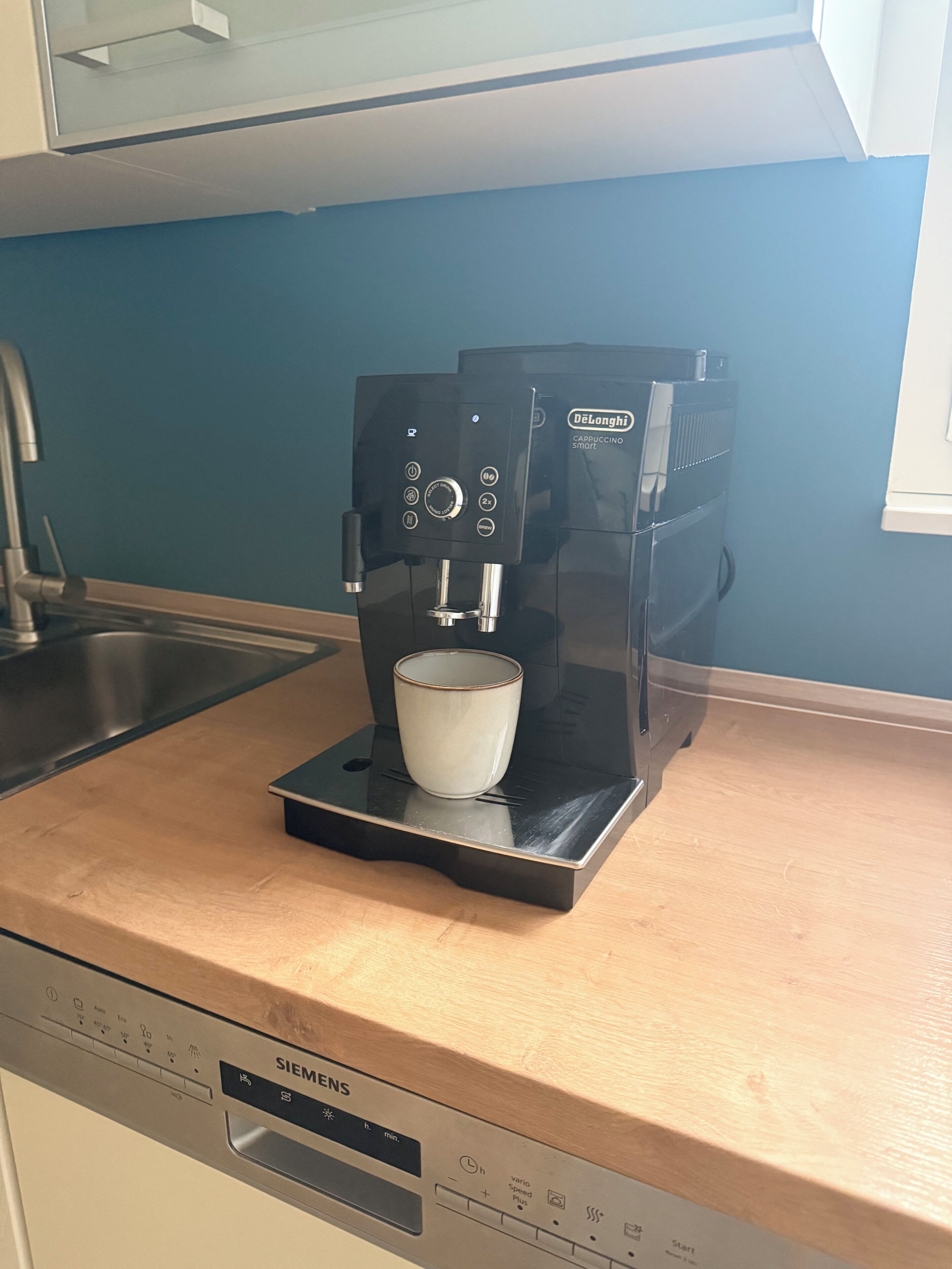 Eine Kaffeemaschine mit einer Tasse auf dem Tablett, darunter ein Siemens Geschirrspüler und eine blaue Wand im Hintergrund.