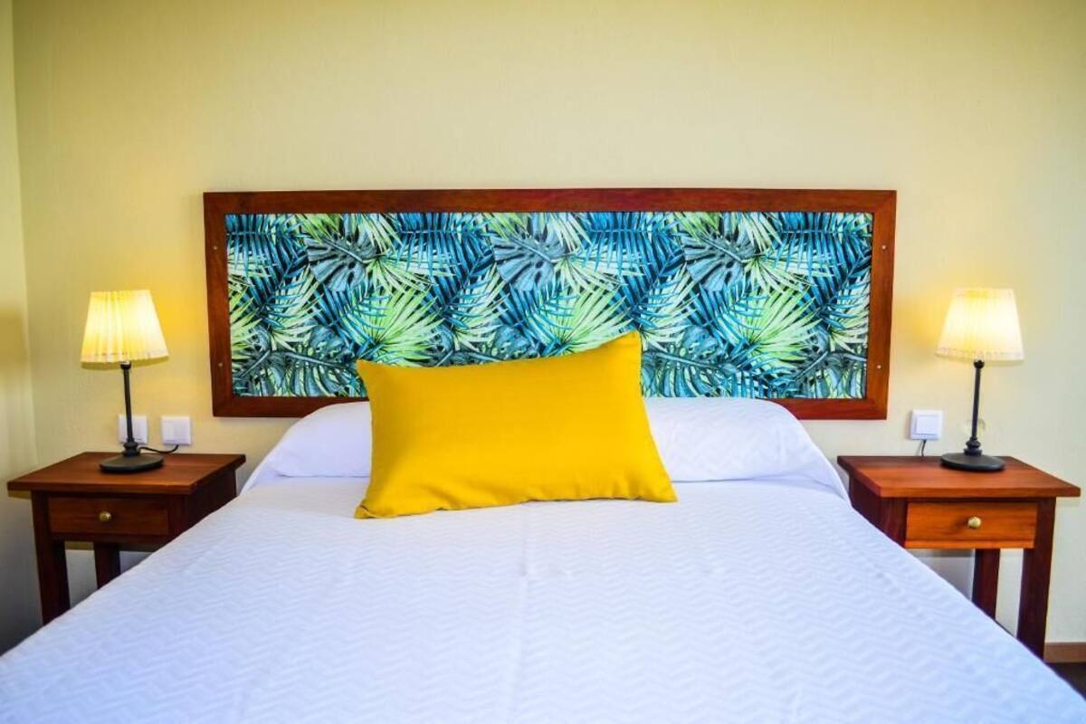 A hotel room with a large bed, two bedside tables with lamps, a bright yellow pillow, and a tropical leaf-patterned headboard.